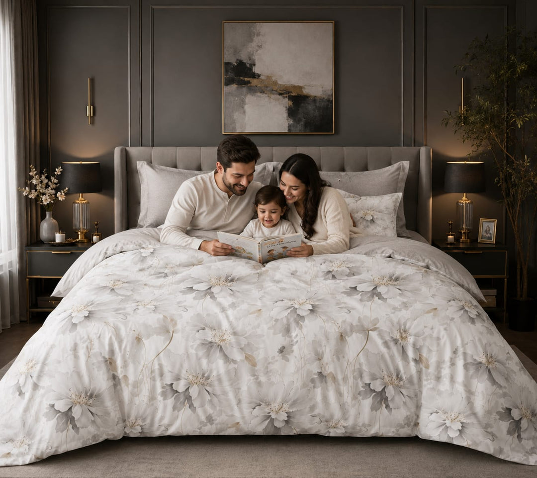 A family of three sits together on a bed in a modern beddings set, reading a book. The bed is covered with a light gray and white floral duvet cover.