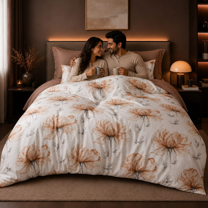 A couple sitting together in bedspread set under a duvet with a large floral print. The bedding has a warm off-white background with large tan and brown flower motifs and subtle gray leaf details.