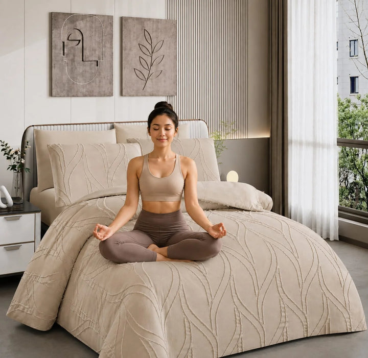 A woman in a beige king size duvet set
 and brown leggings sits cross-legged in meditation pose on a bed with a matching beige quilted comforter and pillow shams in a modern bedroom.
